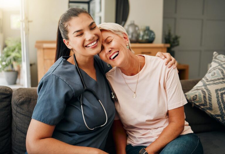 A young female medical caregiver or nursing staff with olive skin tone hugging a senior woman from the side, on the couch in a living room, appearing comfortable and trusting with a supportive, secure bond.