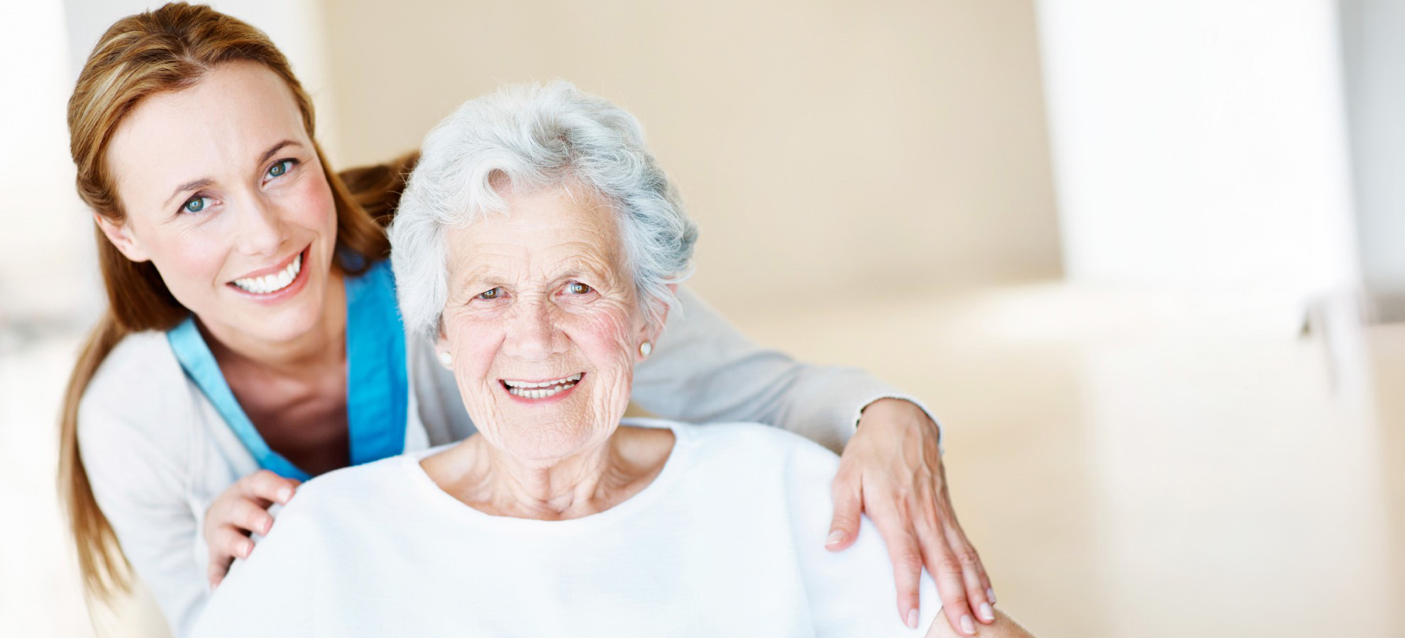 A professional nurse or CNA standing behind an elderly female patient, leaning forward over her shoulder, both smiling.