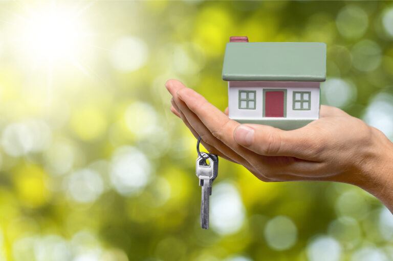 A tiny model house in a Caucasian adult's palm with a single keyring hanging off their ring finger, in front of a blurred lush, green background with bright sunlight.