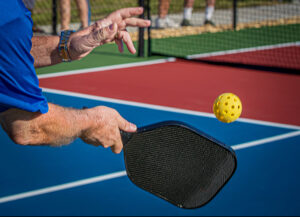 An over-the-shoulder shot of an elderly man hitting a pickleball with a paddle.