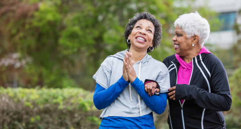 Two African-American women, best friends, hanging out together, walking arm in arm, talking and smiling. The senior woman on the right with white hair is in her 60s. Her friend, who is in her 50s, is looking up at the sky with her hands folded, gesturing a prayer or wishful thought.